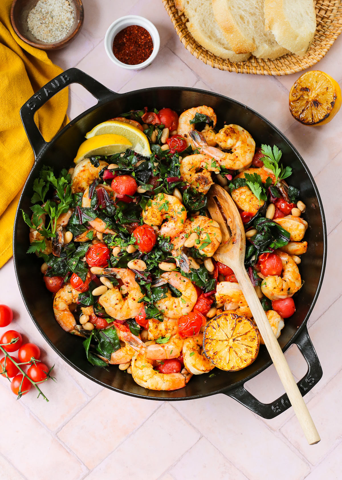 A large braising skillet filled with seared shrimp, white beans, blistered tomatoes, and garlicky greens with lemon wedges for garnish and a wooden serving spoon resting on the side of the skillet.
