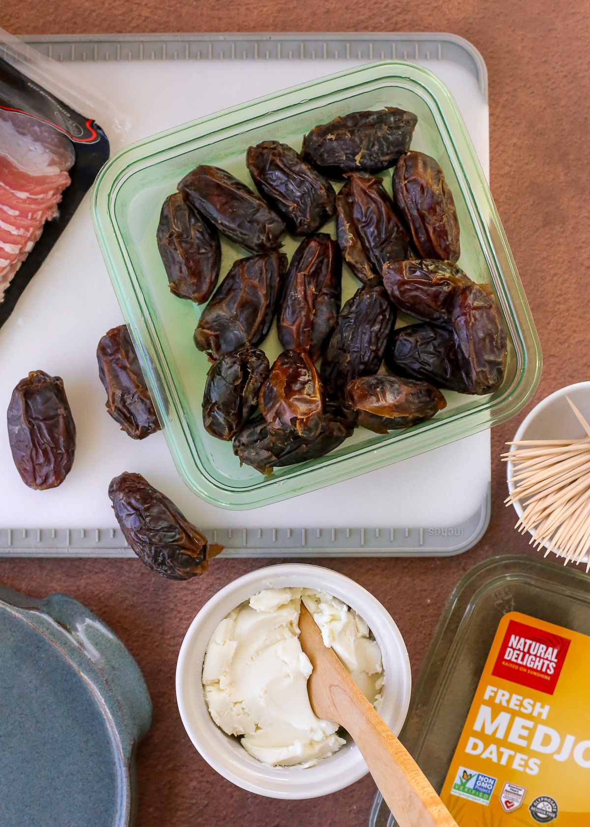 An open container of Medjool dates sitting on a cutting board, with a prep bowl of creamy cheese, a ramekin of toothpicks, and an open package of bacon nearby. 