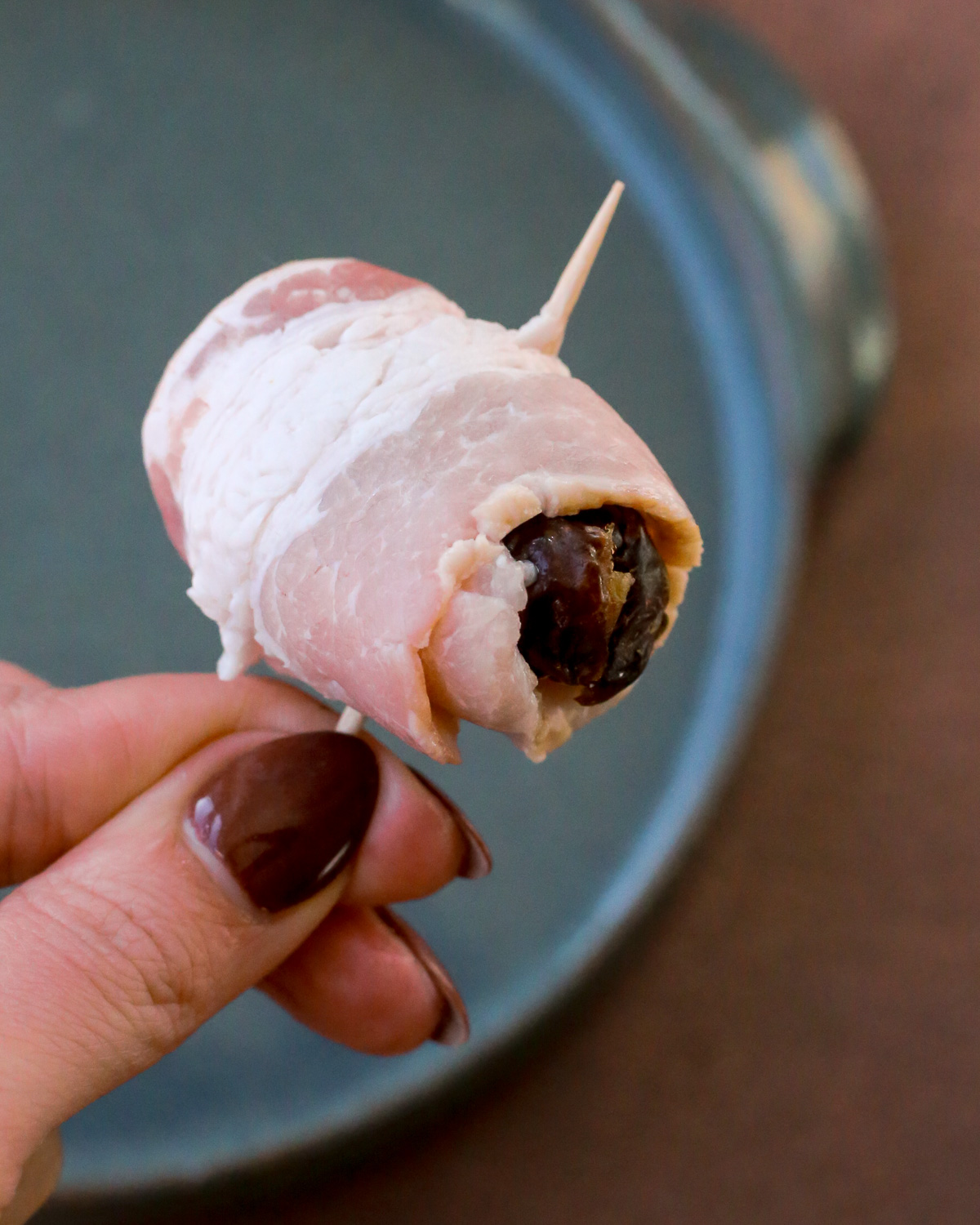 A woman's hand holds a stuffed date wrapped in uncooked bacon, secured with a toothpick, and ready to go into the air fryer. 