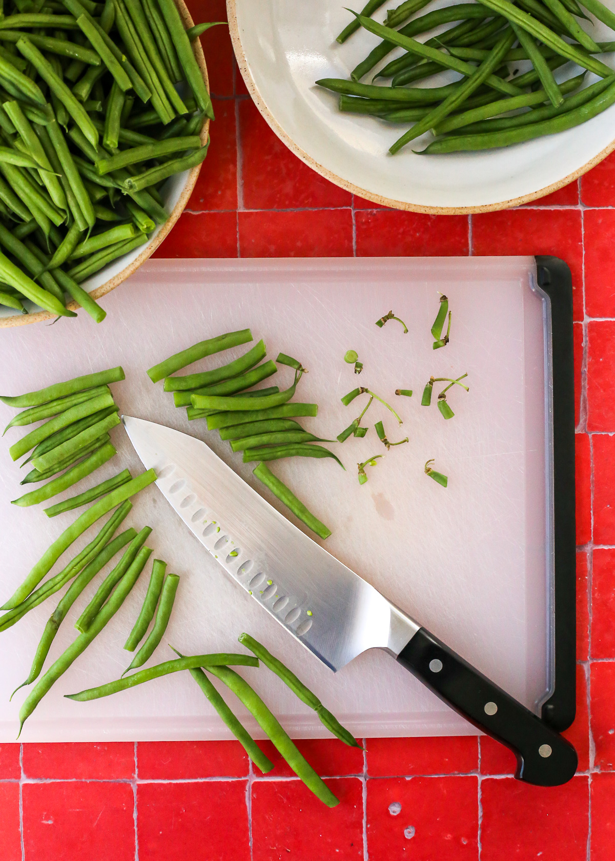 A cutting board and chefs knife with trimmed green beans, indicating how to cut them before blanching.
