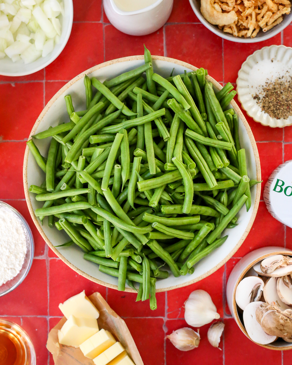 A large bowl of trimmed green beans surrounded by ingredients like diced onions, sliced mushrooms, garlic cloves, cream, butter, and Boursin cheese.
