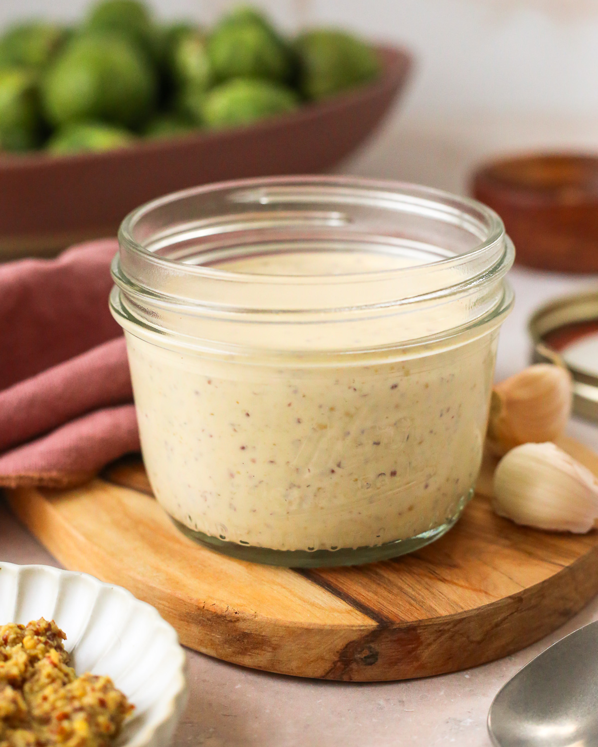 A small glass jar of dijon aioli displayed on a kitchen countertop with brussels sprouts in the background.