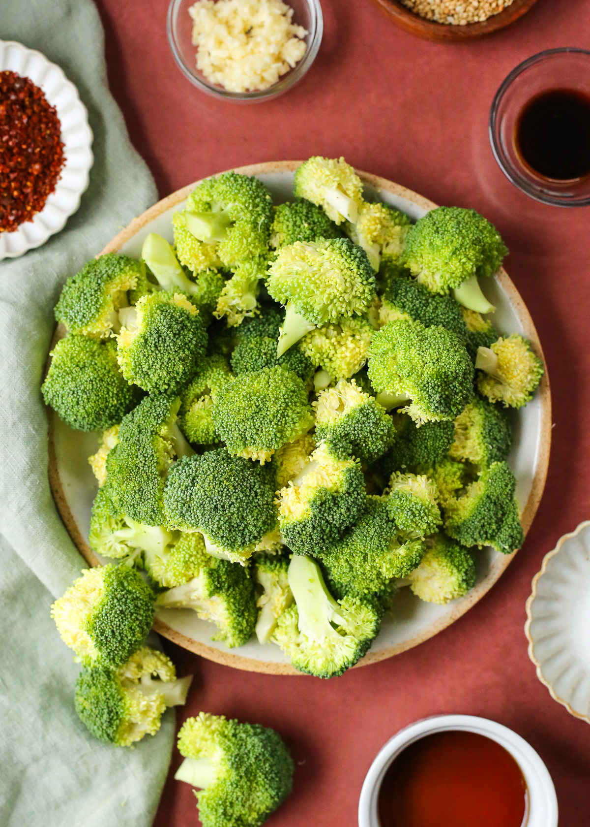 A large plate filled with raw, trimmed broccoli florets surrounded by ingredients for a Korean broccoli side dish. 