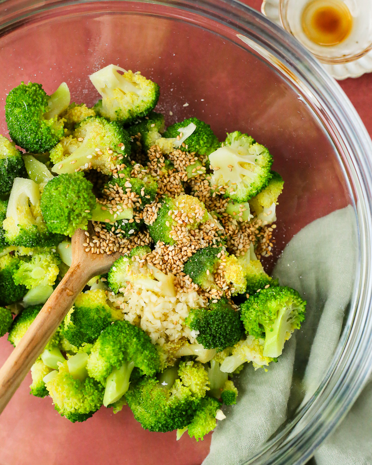 A clear glass mixing bowl containing steamed broccoli dressed with minced garlic, sesame seeds, sesame oil, soy sauce, and sugar. 