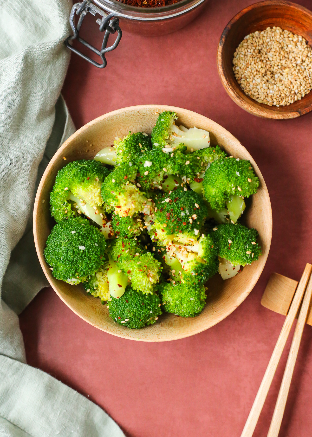 A small wooden bowl containing steamed broccoli seasoned with sesame seeds and pepper flakes, with a pair of wooden chopsticks resting on the side.